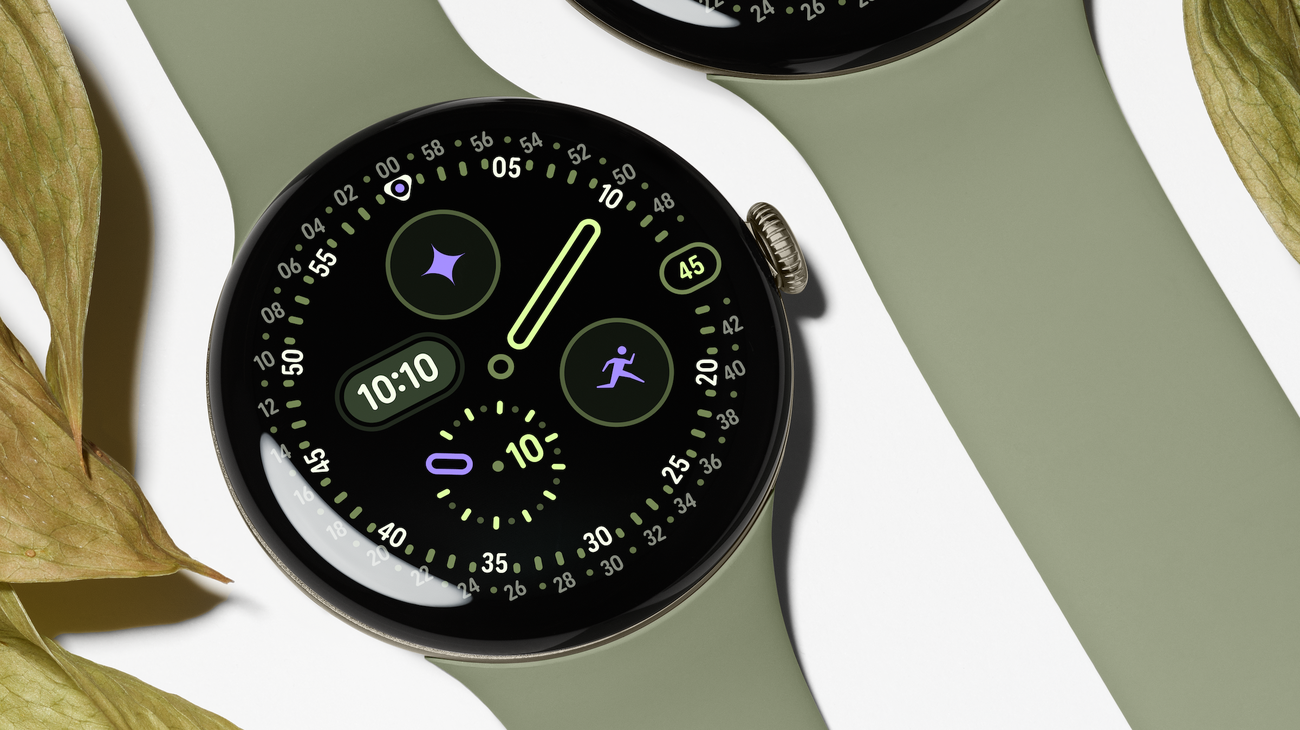 Google Pixel Watch 5 with a round domed display and olive Active Band lying flat beside leaves