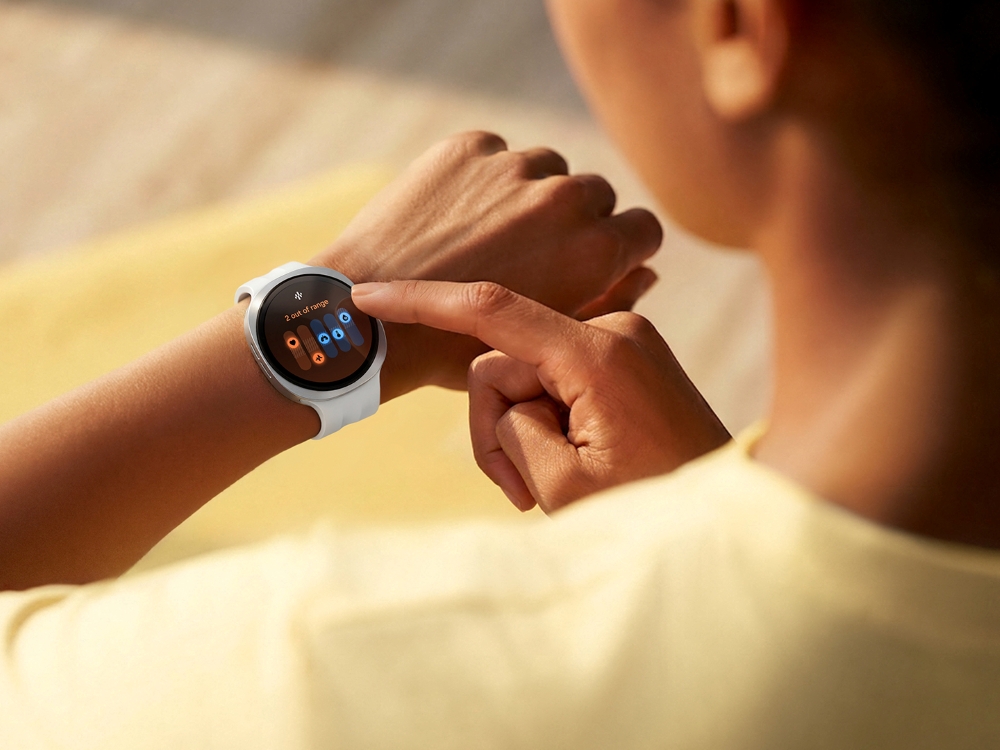 Samsung Galaxy Watch 9 in a light finish worn on the wrist, with the wearer touching the side of the watch